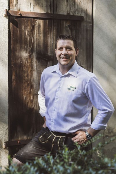 Wirt Raimund Mann, © Niederösterreich Werbung/David Schreiber Mann in Tracht vor Holztür, trägt Hemd mit Logo, lächelt in die Kamera.