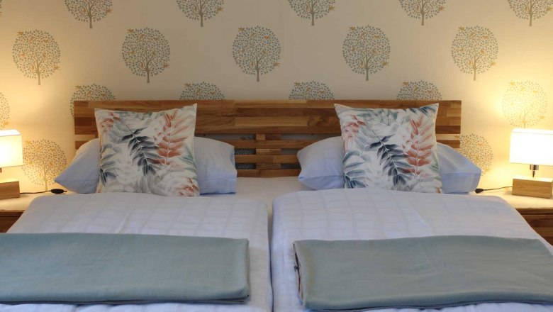 Gemütliche Zimmer, © Magdalena Donhauser Gemütliches Doppelbett mit Holzkopfteil, Decken und Kissen und Nachttischen mit Lampen.