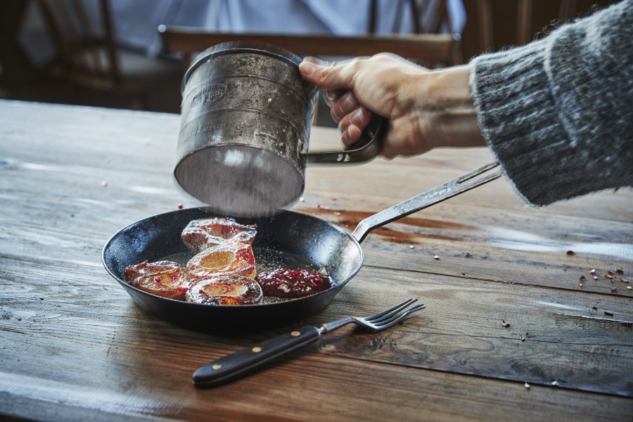 Karamellisierte Apfelringe mit Preiselbeermarmelade, © Niederösterreich Werbung/Andreas Hofer Eine Hand streut Puderzucker über karamellisierte Apfelringe in einer Pfanne auf einem Holztisch.