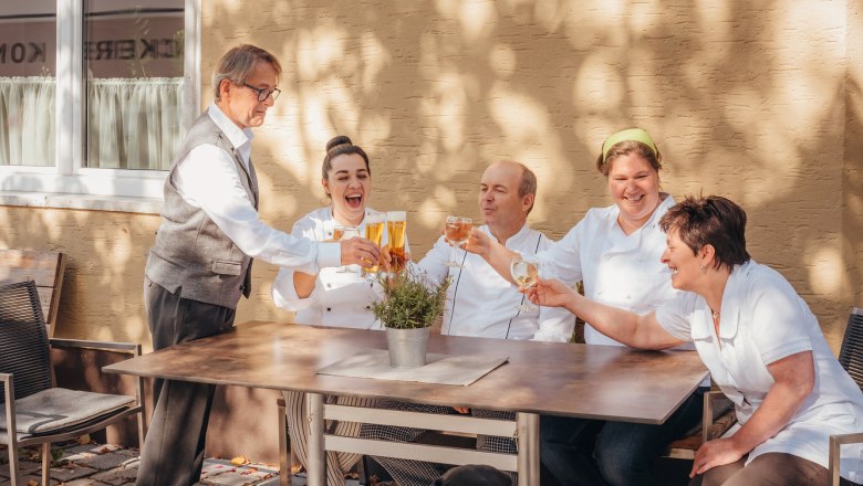 Starkes Wirtshaus-Team, © Niederösterreich Werbung/Daniela Führer Ein Team von fünf Personen sitzt draußen an einem Tisch und stößt mit Getränken an. Ein schwarzer Hund sitzt davor.