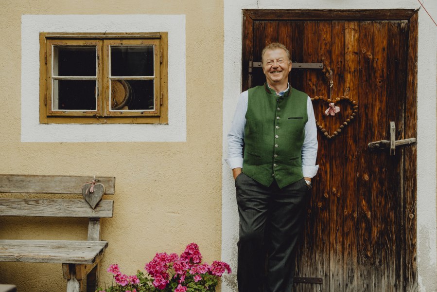 Wirt Anton Schilling, © Niederösterreich Werbung/Rita Newman Ein Mann in traditioneller Kleidung steht lächelnd vor einer Holztür neben einem Fenster und einer Bank mit Blumen.