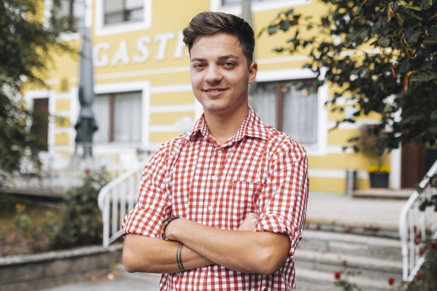 Im Service: Martin Scheiterer, © Niederösterreich Werbung/David Schreiber Ein junger Mann in kariertem Hemd steht lächelnd vor einem gelben Gebäude.