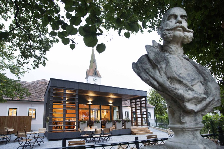 Das FRANZ JOSEPH Wirtshaus am Kaiserplatz, © Astrid Bartl Büste vor modernem Gebäude mit Kirchturm im Hintergrund, umgeben von Bäumen.