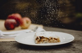 Hausgemachter Apfelstrudel, © Niederösterreich Werbung/Sophie Menegaldo Apfelstrudel mit Puderzucker auf Teller, Äpfel im Hintergrund.