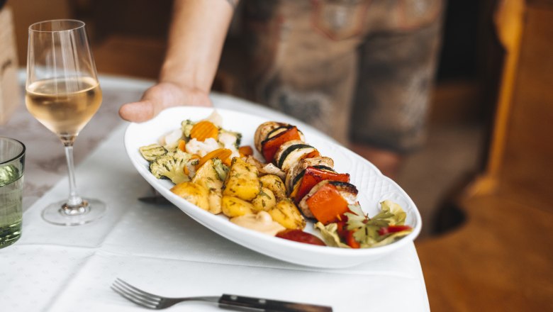 Ein Wirtshausklassiker: Grillspieß, © Niederösterreich Werbung/David Schreiber Ein Teller mit Grillspieß, Gemüse und Kartoffeln wird serviert, daneben ein Glas Weißwein.