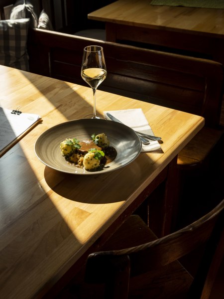 Geschmorter Schweinebauch, Chilikraut, Erdäpfelknödel in knusperiger Schweineschwarte gewälzt, © Niederösterreich Werbung/Michael Reidinger Teller mit Fleisch, Knödeln und Soße, Weinglas auf Holztisch im Sonnenlicht.