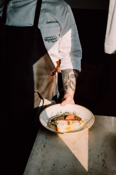 Haubenlokal mit qualitätsvoller österreichischer Küche, © Niederösterreich Werbung/Julius Hirtzberger Koch in Schürze präsentiert ein kunstvoll angerichtetes Gericht auf einem Teller.