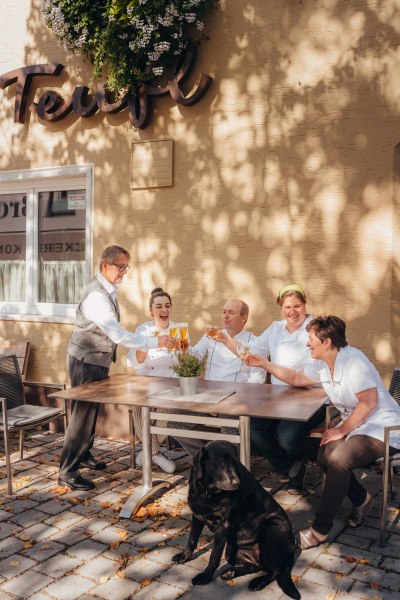 Starkes Wirtshaus-Team, © Niederösterreich Werbung/Daniela Führer Ein Team von fünf Personen sitzt draußen an einem Tisch und stößt mit Getränken an. Ein schwarzer Hund sitzt davor.