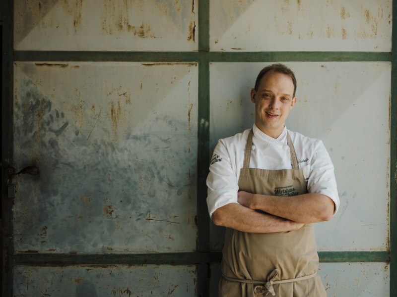 Wirt Patrick Friedrich, © Niederösterreich Werbung/Michael Reidinger Koch in weißer Uniform und Schürze vor einer alten Metalltür.