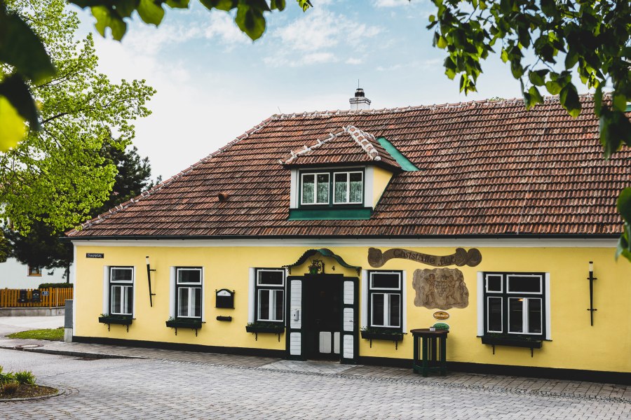 Gaststätte Figl in St. Pölten (Ratzersdorf), © Niederösterreich Werbung/David Schreiber Gelbes Gebäude mit roten Dachziegeln und grünen Fensterläden, umgeben von Bäumen.