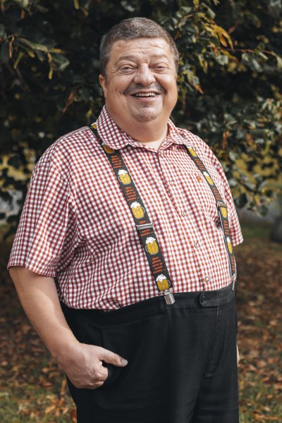 Wirt: Herbert Scheiterer, © Niederösterreich Werbung/David Schreiber Ein Mann in kariertem Hemd und Hosenträgern steht lächelnd vor einem Baum.