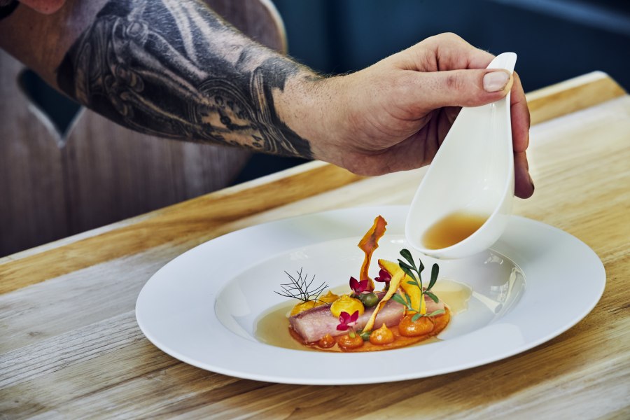 Hier isst das Auge mit, © Niederösterreich Werbung/Andreas Hofer Ein Teller mit kunstvoll angerichtetem Essen, über das eine Hand aus einer kleinen Kanne Sauce gießt.