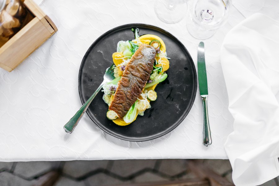 Neubrucker Saiblingsfilet, © Niederösterreich Werbung/Mara Hohla Ein kunstvoll angerichtetes Saiblingsfilet auf einem schwarzen Teller mit Gemüse und Sauce, auf einem weißen Tisch.