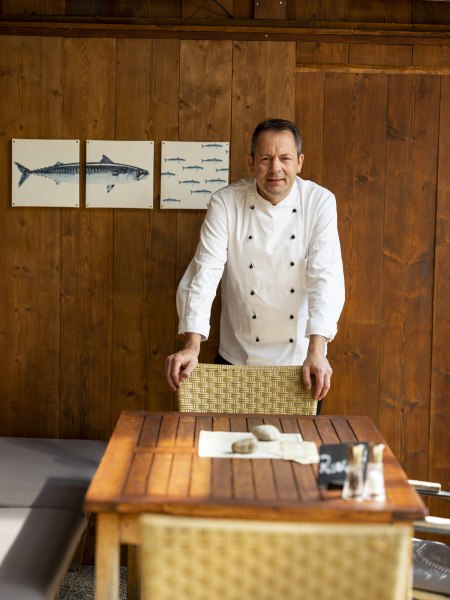 Wirt Martin Weiler, © Niederösterreich Werbung/Michael Reidinger Ein Mann in Kochkleidung steht an einem Holztisch vor einer Holzwand mit Fischbildern.
