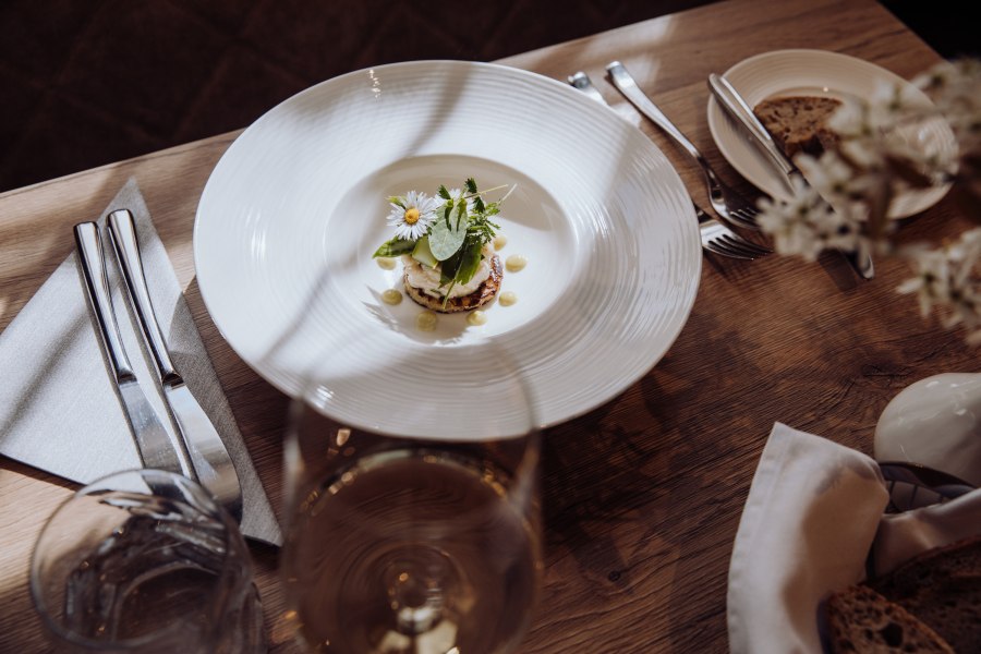 Tartar von der Räucherforelle auf Erdäpfelpuffer, © Niederösterreich Werbung/thecreatingclick.com Ein Teller mit Tartar von Räucherforelle auf einem Erdäpfelpuffer, garniert mit Kräutern und Blüten, auf einem Holztisch mit Besteck und einem Glas Wein.