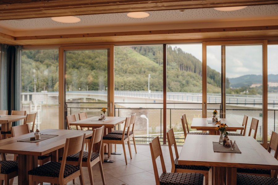 Die Donau und die Stadt Grein immer im Blick, © Niederösterreich Werbung/Daniela Führer Innenansicht eines Gasthofes mit großen Fenstern und Blick auf eine Brücke und bewaldete Hügel.