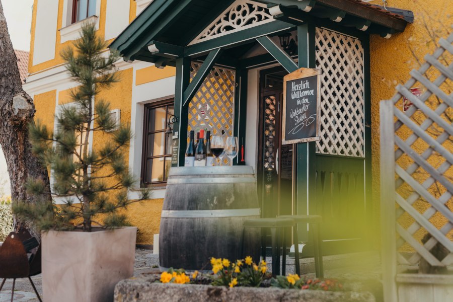 Wirtshaus im ersten Faulenzerhotel Österreichs, © Niederösterreich Werbung/Daniela Führer Eingang eines Wirtshauses mit Weinfass, Gläsern und Blumen.