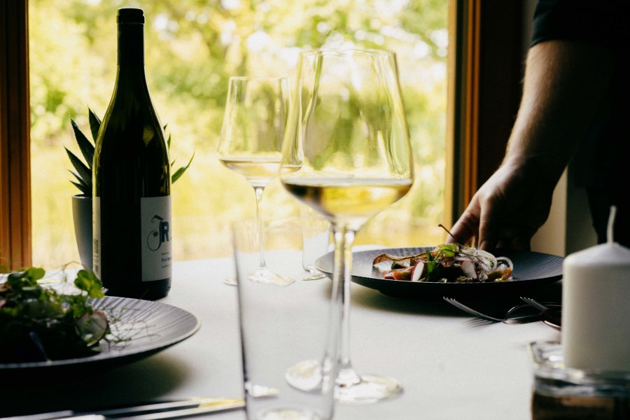 Restaurant im Weingut Johnneshof Reinisch, © Julius Hirtzberger Ein gedeckter Tisch mit Weinflasche, Weingläsern und einem Teller mit Essen in einem Restaurant.