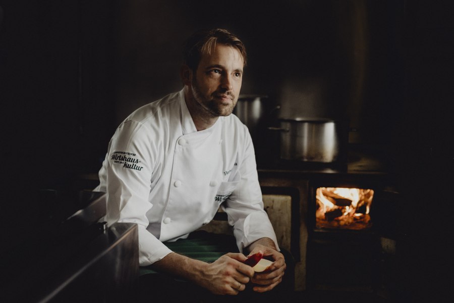 Herbert Bonka`s Schweinsbraten kommt aus dem 100-jährigen Holzofen, © Niederösterreich Werbung/Sophie Menegaldo Koch in weißer Uniform vor einem traditionellen Herd mit Feuer.