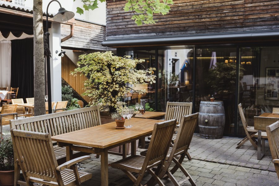 Grüner Hofgarten, © Niederösterreich Werbung/David Schreiber Ein gemütlicher Innenhof mit Holzmöbeln, Pflanzen und einem Weinfass, umgeben von Gebäuden mit Holzverkleidung.