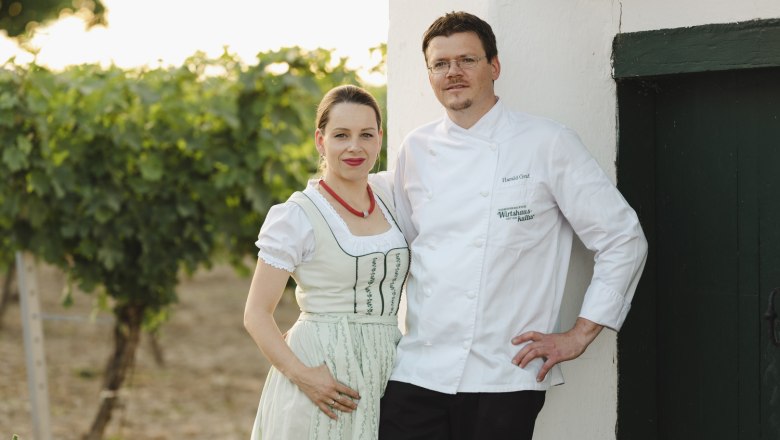 Wirtsleute Daniela und Harald Graf, © Niederösterreich Werbung/Michael Reidinger Ein Paar in traditioneller Kleidung steht vor einem weißen Gebäude neben einem Weinberg.
