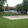 Theater- und Feriendorf Königsleitn, © Theater- und Feriendorf Königsleitn Menschen spielen Volleyball auf einem Sandplatz im Freien, umgeben von Bäumen und einem Gebäude im Hintergrund.