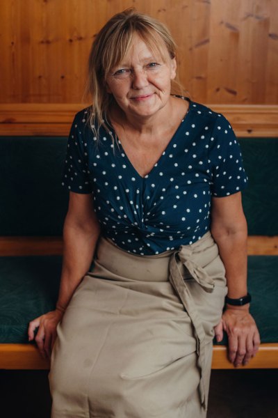 Wirtin Gabriele Hader, © Niederösterreich Werbung/Daniela Führer Frau in gepunkteter Bluse und Schürze sitzt auf einer Holzbank.