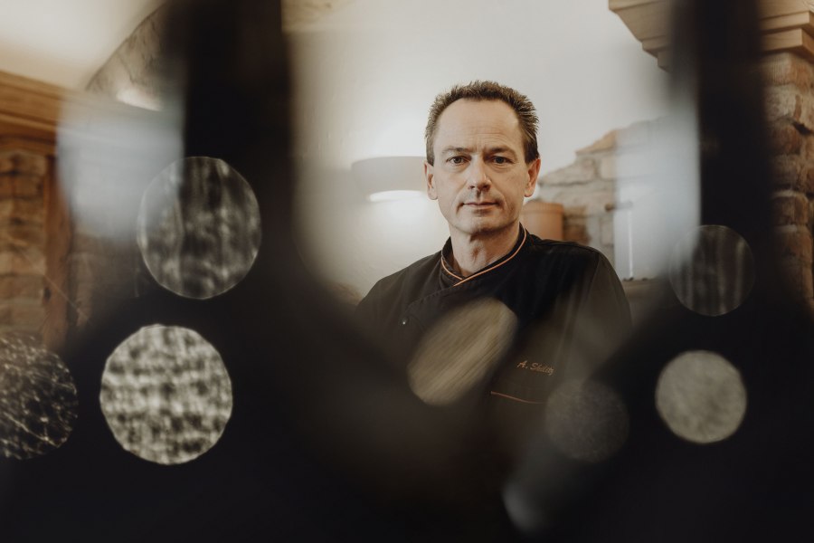 Wirt Alfred Skilitz, © Niederösterreich Werbung/Sophie Menegaldo Ein Mann in schwarzer Kochkleidung steht in einem rustikalen Raum, umgeben von unscharfen Lichteffekten.