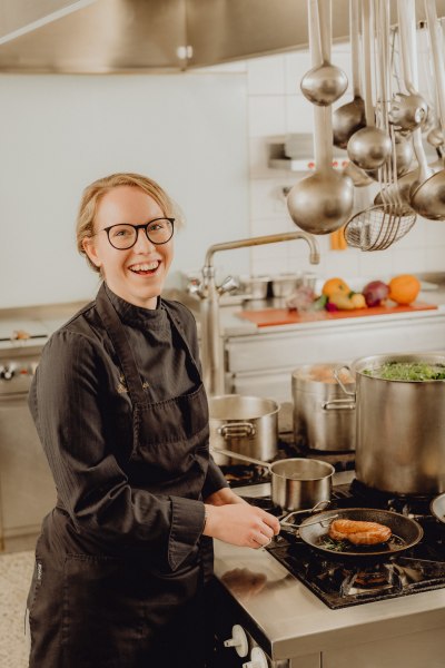 Köchin Maria Buchleitner, © Niederösterreich Werbung/Klaus Engelmayer Eine Köchin in schwarzer Uniform steht lächelnd in einer professionellen Küche und brät ein Stück Fleisch in einer Pfanne.