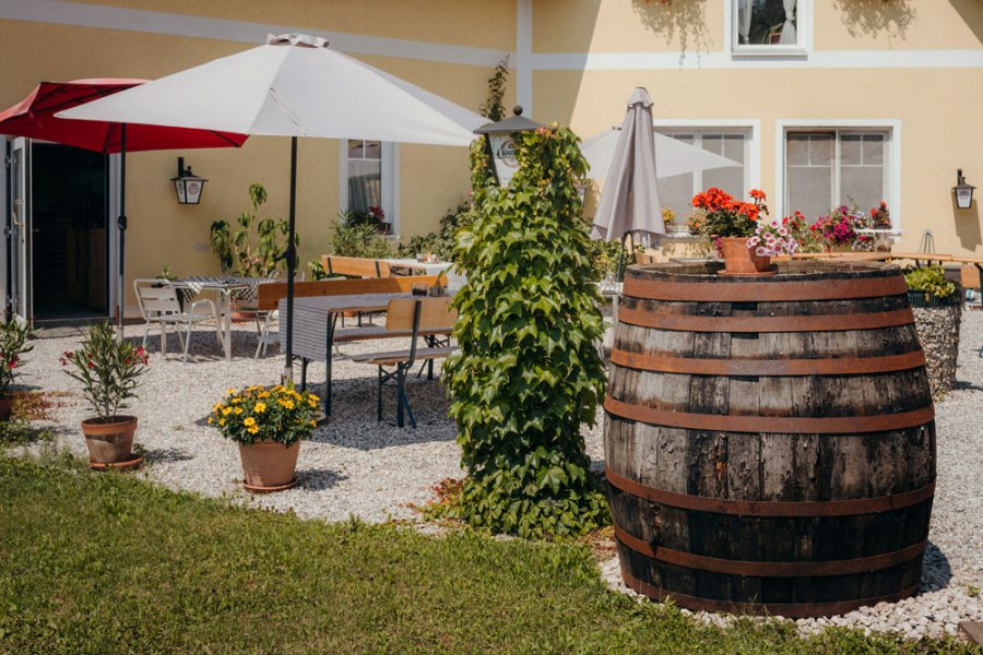 Platz nehmen im grünen Gastgarten, © Niederösterreich Werbung/Daniela Führer Ein gemütlicher Gastgarten mit Sonnenschirmen, Holztischen und einem großen Holzfass, umgeben von Pflanzen und Blumen.