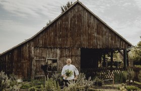Wirtshaus mit Weinviertler Kochkursen, © Niederösterreich Werbung/Sophie Menegaldo Ein Koch steht vor einer alten Holzhütte mit einem Kräuterstrauß in der Hand.
