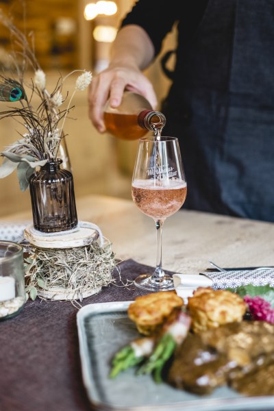 Mit Steinpilzen gefüllter Rostbraten, © Niederösterreich Werbung/David Schreiber Person schenkt roséfarbenen Wein in ein Glas ein, daneben ein Teller mit Essen und eine Vase mit Trockenblumen.