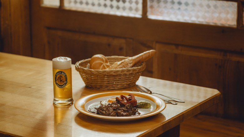 Zum Fiakergulasch passt das helle Fiakerbräu, © Niederösterreich Werbung/Rita Newman Ein Teller mit Fiakergulasch, Spiegelei und Gurke, daneben ein Glas Bier und ein Brotkorb auf einem Holztisch.