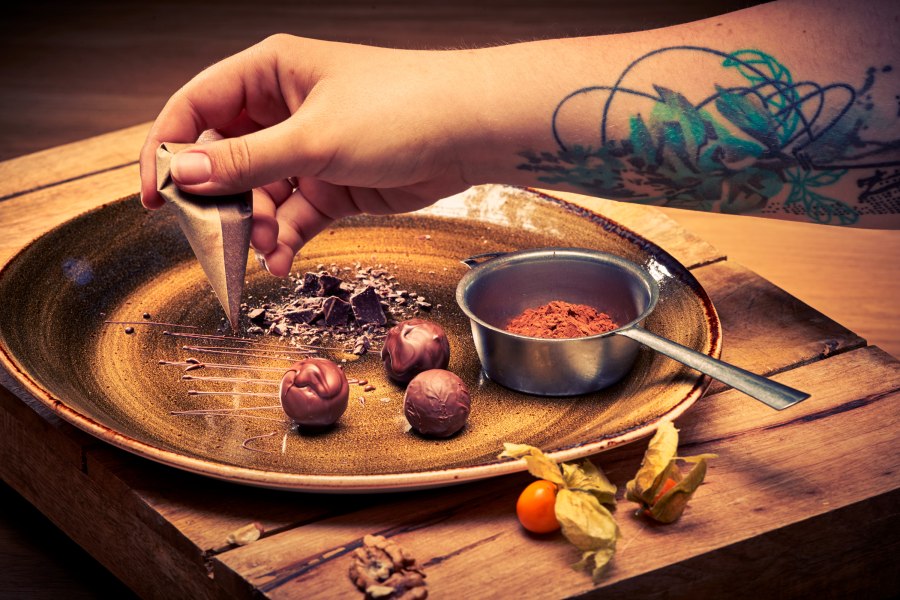 Wirtshaus-Patisserie, © Niederösterreich Werbung/Andreas Hofer Nahaufnahme einer Hand mit Tätowierung, die Schokolade auf einem Teller dekoriert.