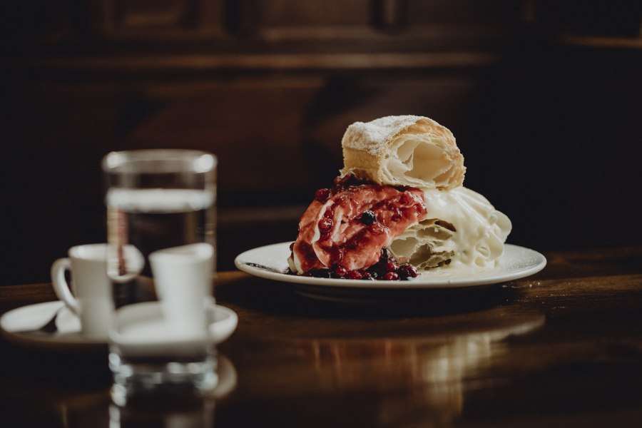 Klostercremeschnitte, © Niederösterreich Werbung/Sophie Menegaldo Ein Teller mit einer Klostercremeschnitte, garniert mit roten Beeren, steht auf einem Holztisch. Im Hintergrund sind ein Glas Wasser und zwei Espressotassen zu sehen.
