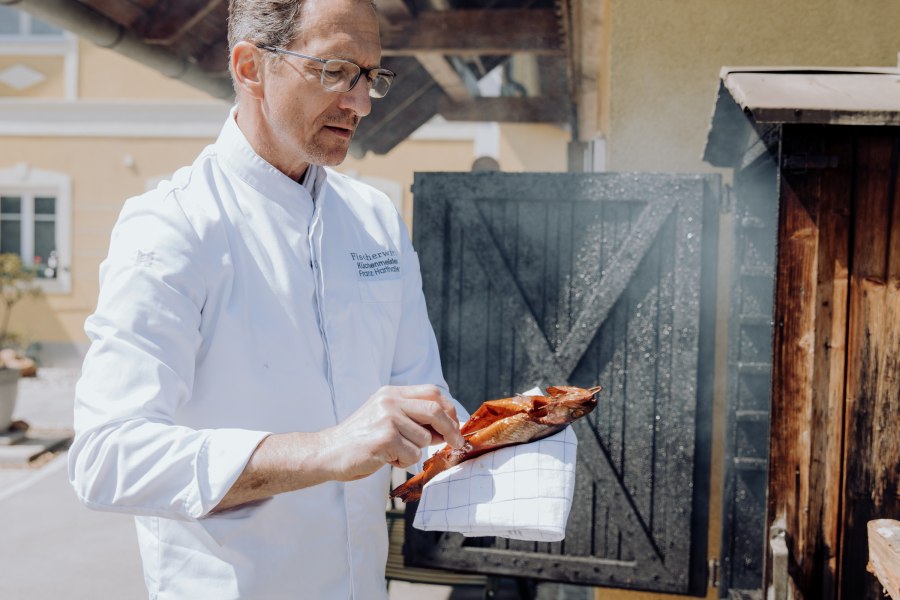 Wirt Franz Harthaller räuchert seine Forellen selbst, © Niederösterreich Werbung/thecreatingclick.com Ein Mann in weißem Kittel hält eine geräucherte Forelle vor einem Räucherofen.