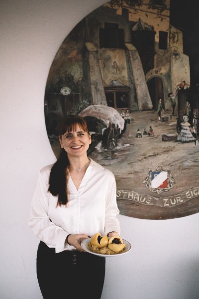 Wirtin Alexandra Turek, © Niederösterreich Werbung/Mara Hohla Frau in weißer Bluse hält Teller mit Knödeln vor einem Wandgemälde.