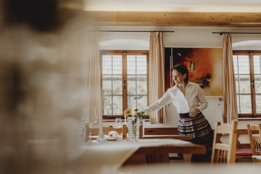 Rundum stimmiges Ambiente, © Niederösterreich Werbung/Sophie Menegaldo Eine Frau in traditioneller Kleidung richtet einen Tisch in einem gemütlichen Raum mit Holzmöbeln und großen Fenstern her.