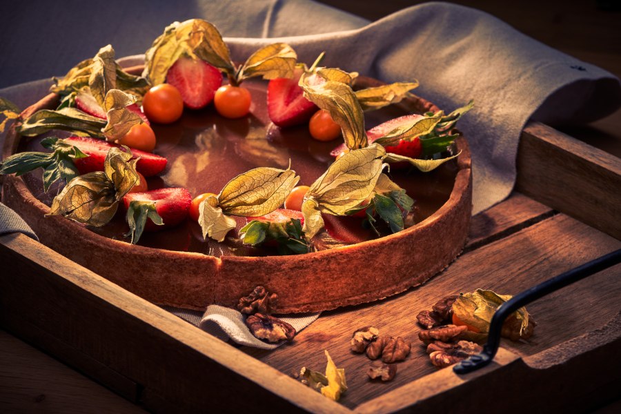 Tortenkreationen, © Niederösterreich Werbung/Andreas Hofer Eine Torte mit Schokoladenglasur, dekoriert mit Erdbeeren und Physalis, auf einem Holztablett.