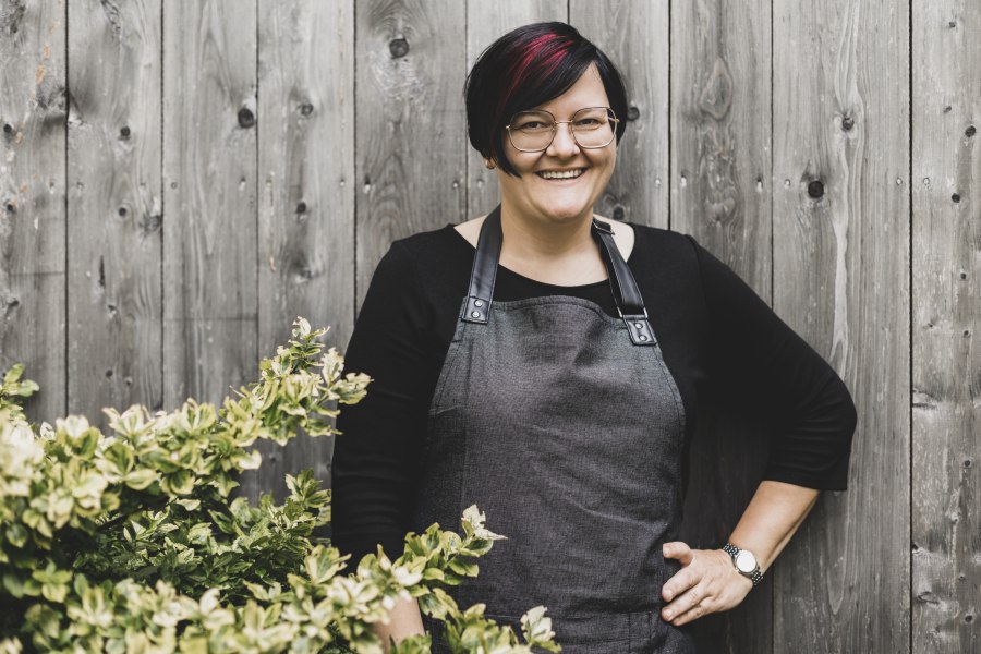 Wirtin Petra Haselsteiner, © Niederösterreich Werbung/David Schreiber Eine Frau mit Brille und Schürze steht lächelnd vor einer Holzwand neben einem Busch.