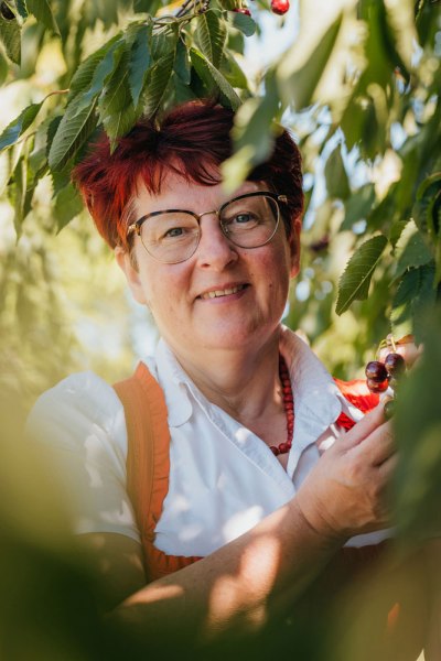 Wirtin Martina Krenn, © Niederösterreich Werbung/Daniela Führer Frau mit Brille und roten Haaren steht unter einem Kirschbaum.