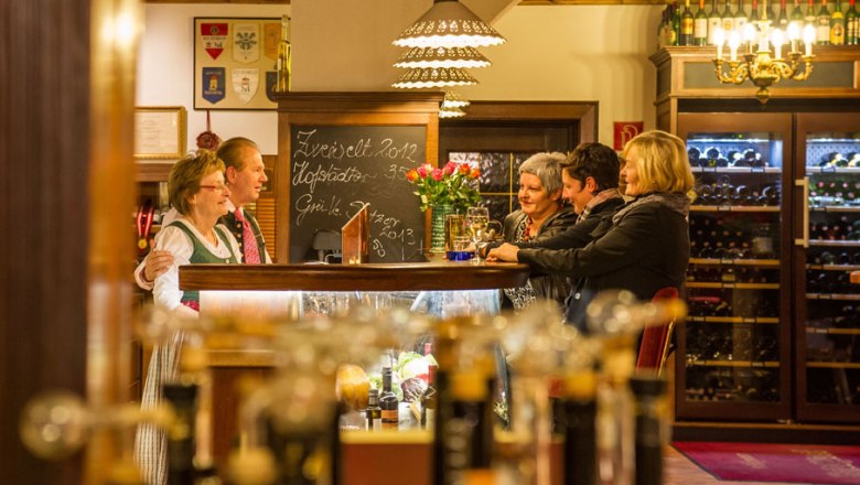 Hönigwirt - Gaststube, © Rita Newman Menschen in einer gemütlichen Gaststube mit Weinflaschen im Hintergrund.