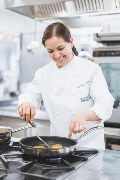 Birgit Heissenberger, © Niederösterreich Werbung/David Schreiber Eine Köchin in weißer Uniform brät Essen in einer Pfanne in einer professionellen Küche.