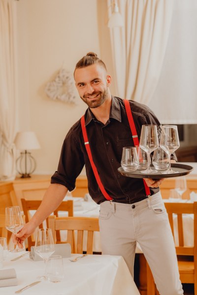 Umsorgt die Gäste: Daniel Franke, © Niederösterreich Werbung/Klaus Engelmayer Ein Kellner mit roten Hosenträgern serviert Gläser in einem Restaurant.