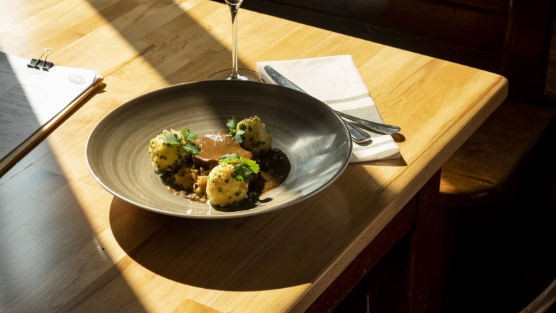 Geschmorter Schweinebauch, Chilikraut, Erdäpfelknödel in knusperiger Schweineschwarte gewälzt, © Niederösterreich Werbung/Michael Reidinger Teller mit Fleisch, Knödeln und Soße, Weinglas auf Holztisch im Sonnenlicht.