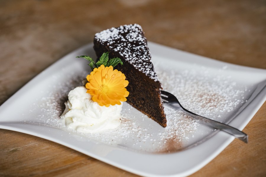 Haustorten mit Schlag, © Niederösterreich Werbung/David Schreiber Ein Stück Schokoladenkuchen mit Puderzucker, Sahne und einer gelben Blume auf einem Teller.