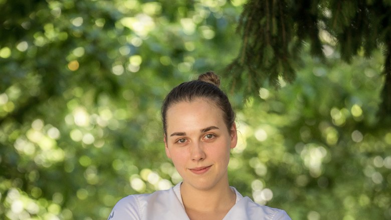 Küchenchefin Victoria Kuben, © Stephan Mussil Eine Frau in weißem Hemd steht mit verschränkten Armen vor einem unscharfen, grünen Hintergrund.