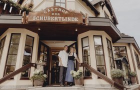 „Zur Schubertlinde“ im Schneebergland, © Niederösterreich Werbung/Rita Newman Eingang des Gasthofs Zur Schubertlinde mit zwei Personen in traditioneller Kleidung.