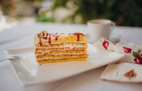 Hagebuttentorte, © Niederösterreich Werbung/Daniela Führer Ein Stück Schichttorte mit Sahne und Karamell auf einem weißen Teller, daneben eine Tasse Kaffee und eine Serviette mit Fruchtmuster.