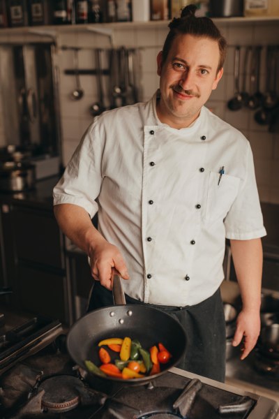 Koch Mathias Scharf, © Niederösterreich Werbung/Daniela Führer Ein Koch in weißer Uniform hält eine Pfanne mit buntem Gemüse in einer Küche.